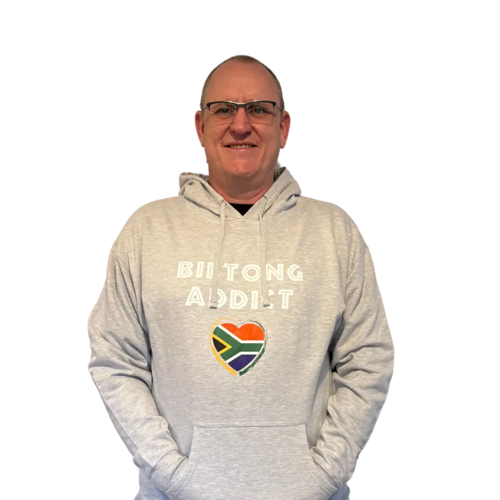 Man wearing a gray Biltong Addict hoodie, smiling and pointing at the design featuring bold text and a South African flag heart.