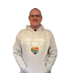 Man wearing a gray Biltong Addict hoodie, smiling and pointing at the design featuring bold text and a South African flag heart.