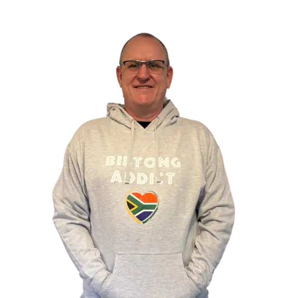 Man wearing a gray Biltong Addict hoodie, smiling and pointing at the design featuring bold text and a South African flag heart.