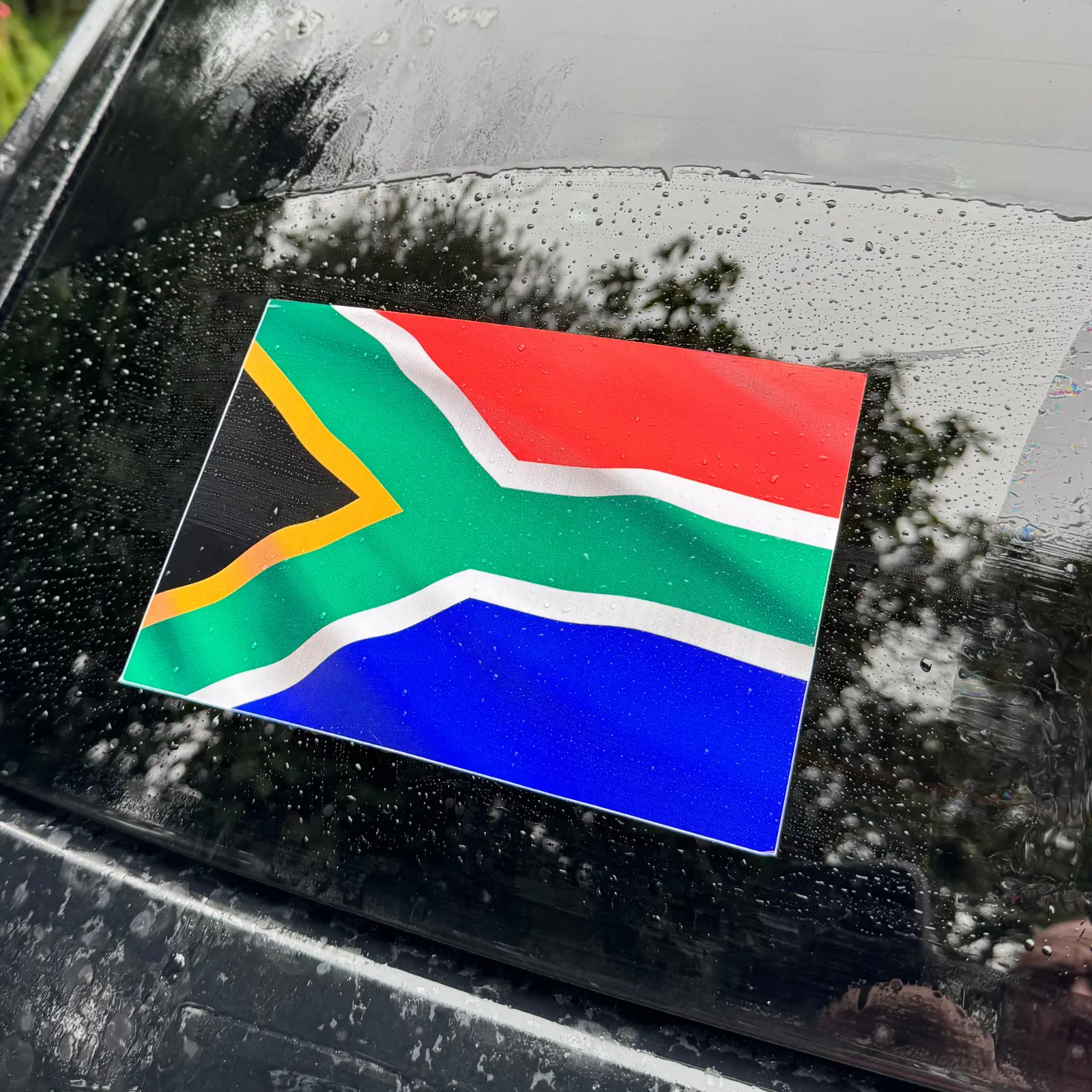 A premium vinyl sticker featuring the South African flag with a waving effect, applied to a car window with raindrops visible.