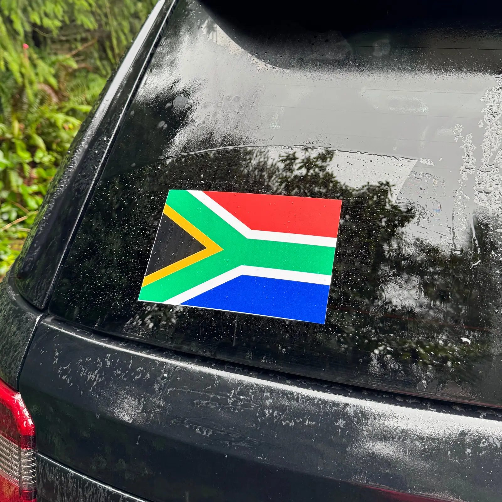 A rectangular South African flag sticker applied to the back window of a black SUV with raindrops on the glass.