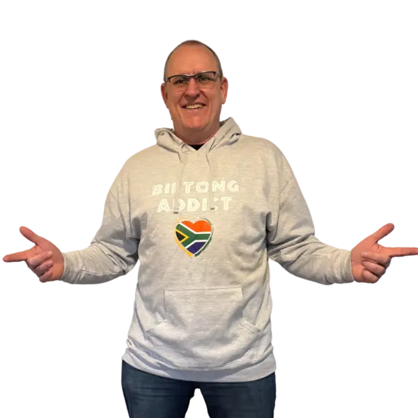 Man wearing a gray Biltong Addict hoodie, smiling and pointing at the design featuring bold text and a South African flag heart.